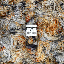 Load image into Gallery viewer, Grey with black and copper speckles yarn with a label on top of a pile of yarn.