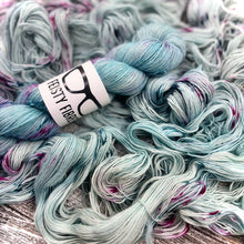 Load image into Gallery viewer, A skein of light teal blue merino linen yarn with  speckles of pink and purple a label that reads &#39;Feisty Fibres&#39;, surrounded by unspooled yarn.
